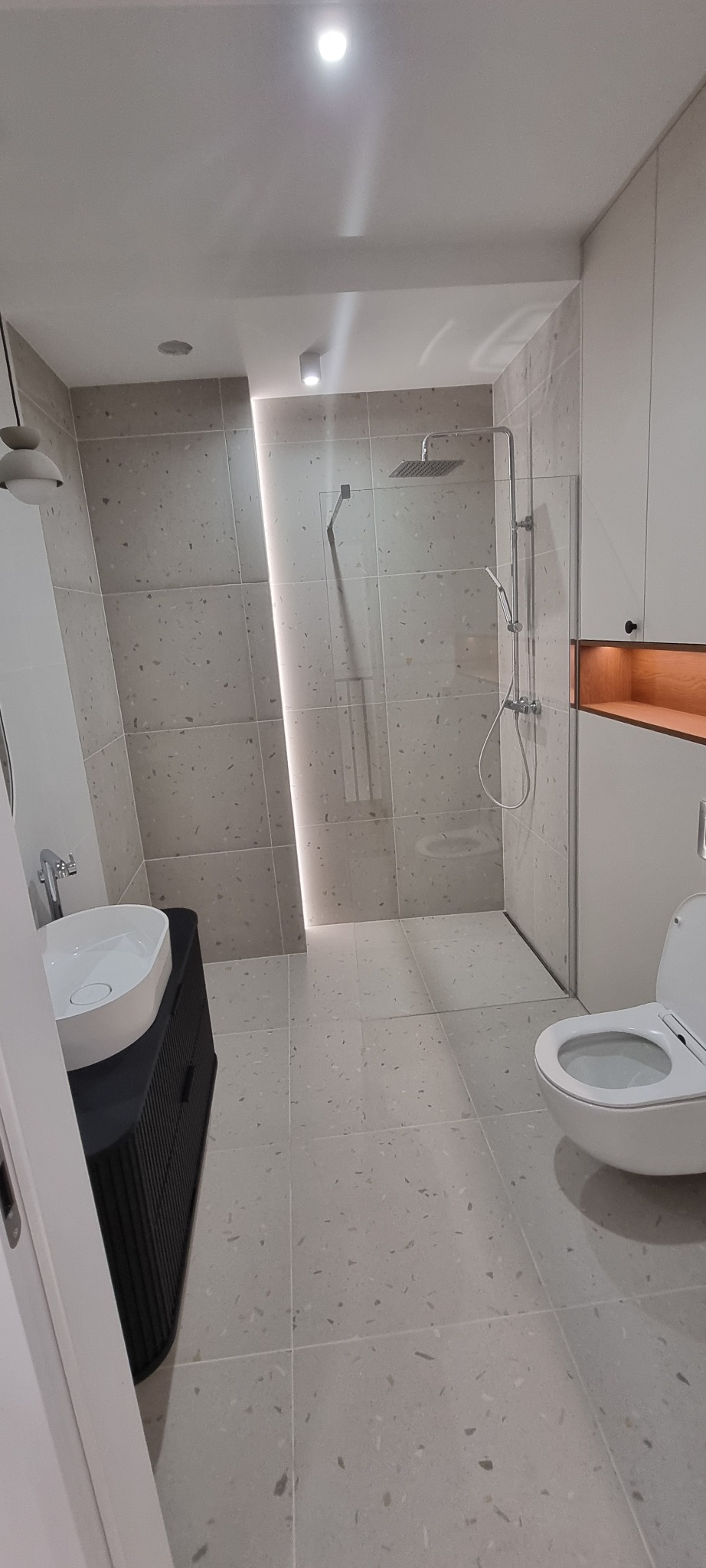 Nowoczesna łazienka z kabiną prysznicową typu walk-in, umywalką nablatową na szafce z lamelami i podświetleniem LED. Jasne płytki imitujące lastryko.