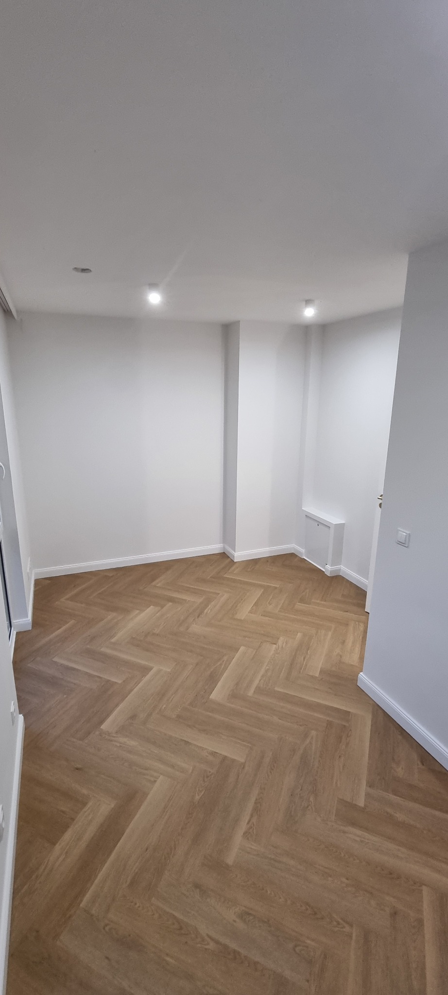 Jasne, puste pomieszczenie z drewnianą podłogą ułożoną w jodełkę. Ściany pomalowane na biało, widoczne listwy przypodłogowe i oświetlenie punktowe.