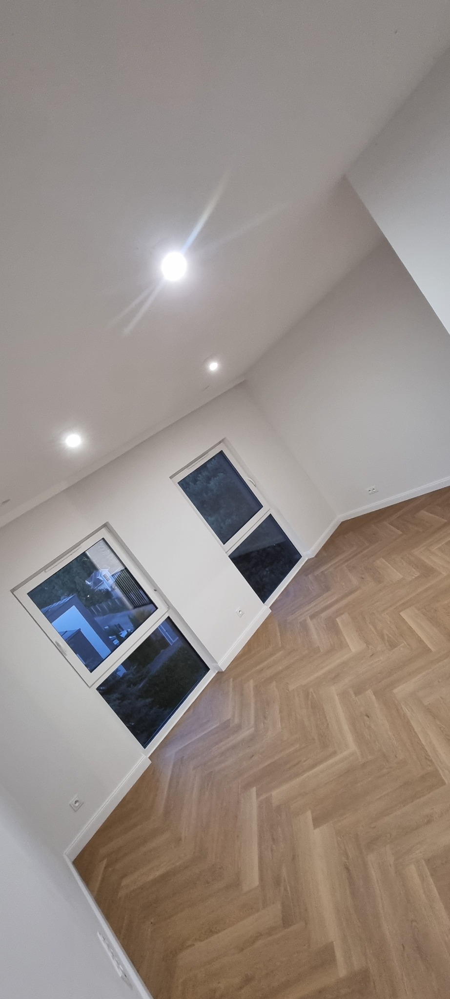 Jasne panele ułożone w jodełkę w nowym, pustym pokoju z dwoma oknami i białymi ścianami. Widoczne gniazdka elektryczne.