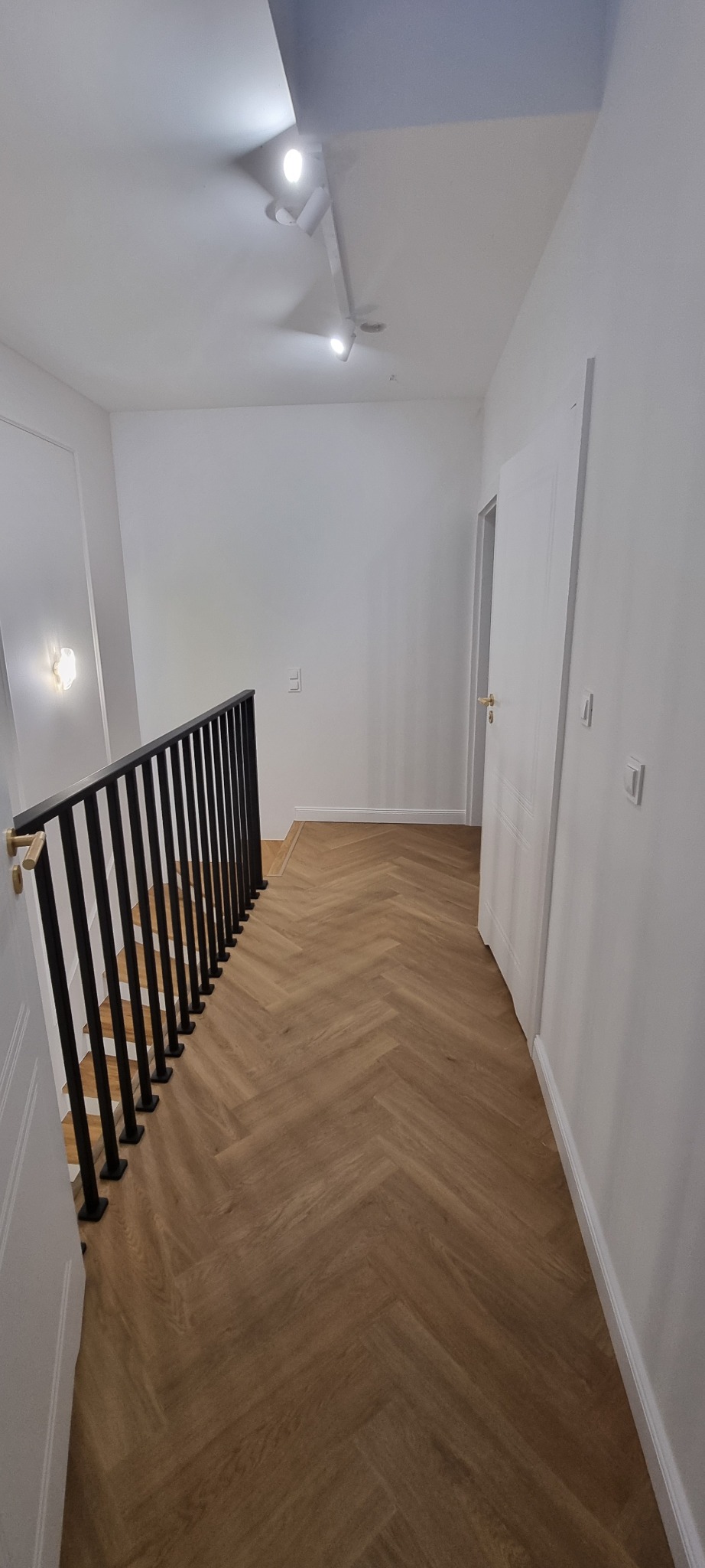 Nowoczesne wnętrze: parkiet w jodełkę, czarna balustrada, białe ściany i drzwi z delikatnym oświetleniem punktowym. Minimalistyczny design w jasnej tonacji.