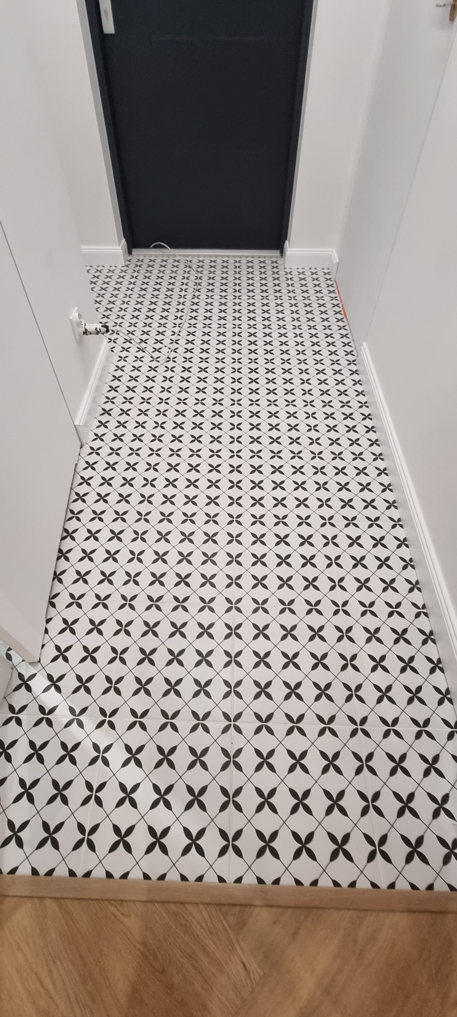 Wzór w czarno-białe płytki podłogowe, ułożone w korytarzu z ciemnymi drzwiami. W tle widoczny fragment drewnianej podłogi. Minimalistyczny design.