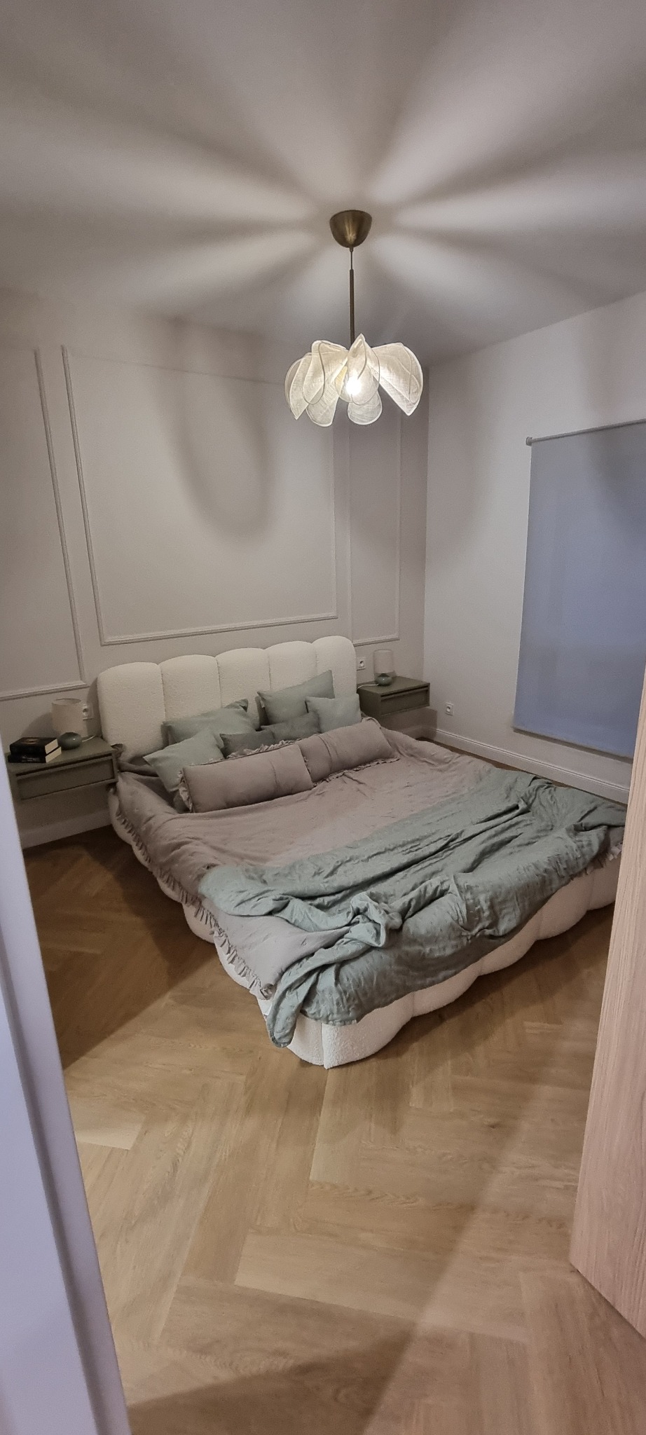 Elegancka sypialnia z parkietem w jodełkę, łóżkiem z miękkim zagłówkiem i designerską lampą w kształcie kwiatu. Minimalistyczne szafki nocne i stonowana kolorystyka.