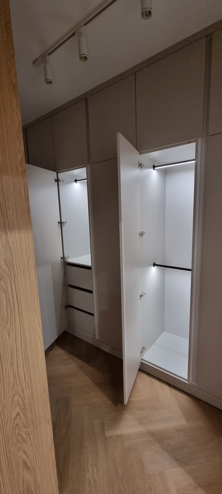 Nowoczesna zabudowa szafy wnękowej z oświetleniem LED i otwartymi drzwiami, ukazująca szuflady i drążki na ubrania. Parkiet w jodełkę na podłodze.