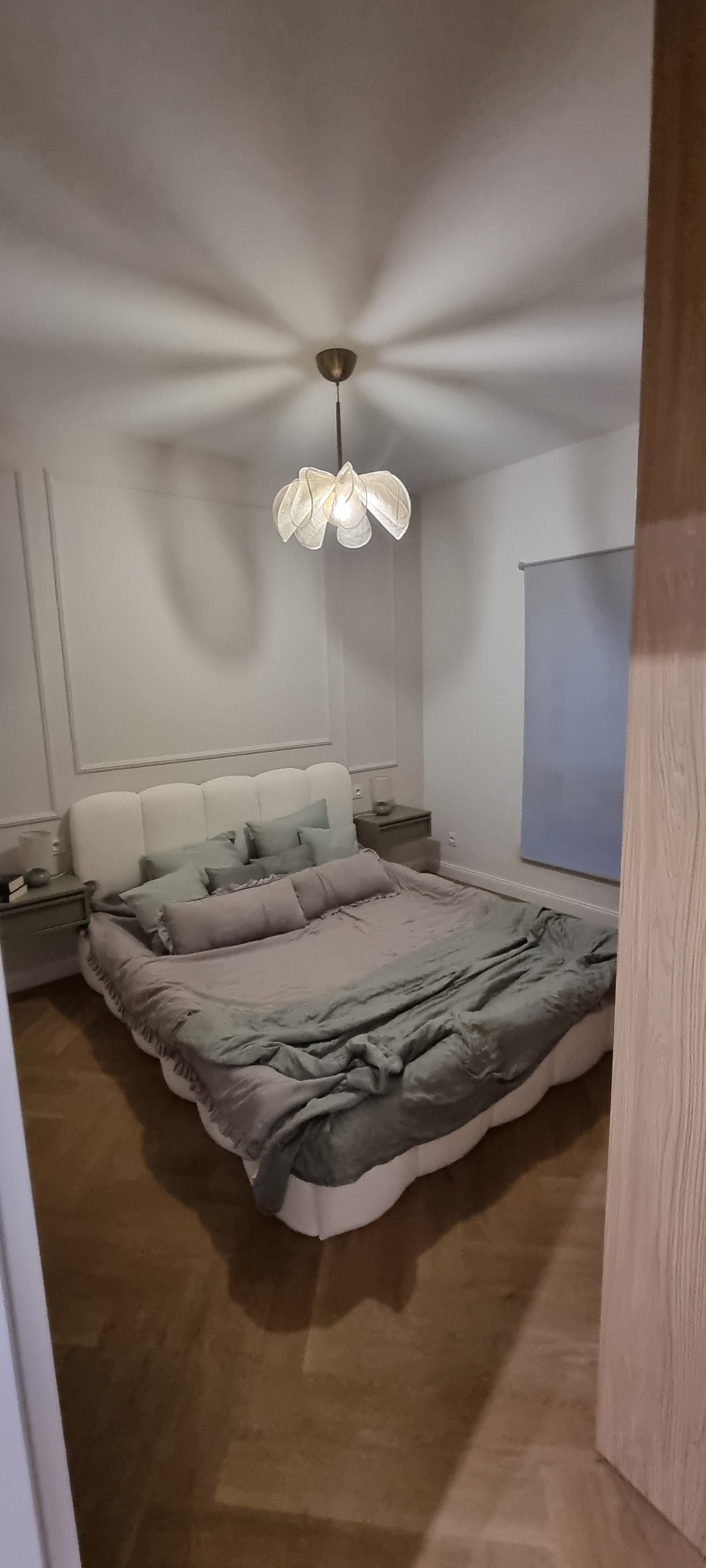 Elegancka sypialnia z dużym łóżkiem, lampą w kształcie kwiatu i szarą pościelą. Ściany zdobione sztukaterią. Parkiet na podłodze.