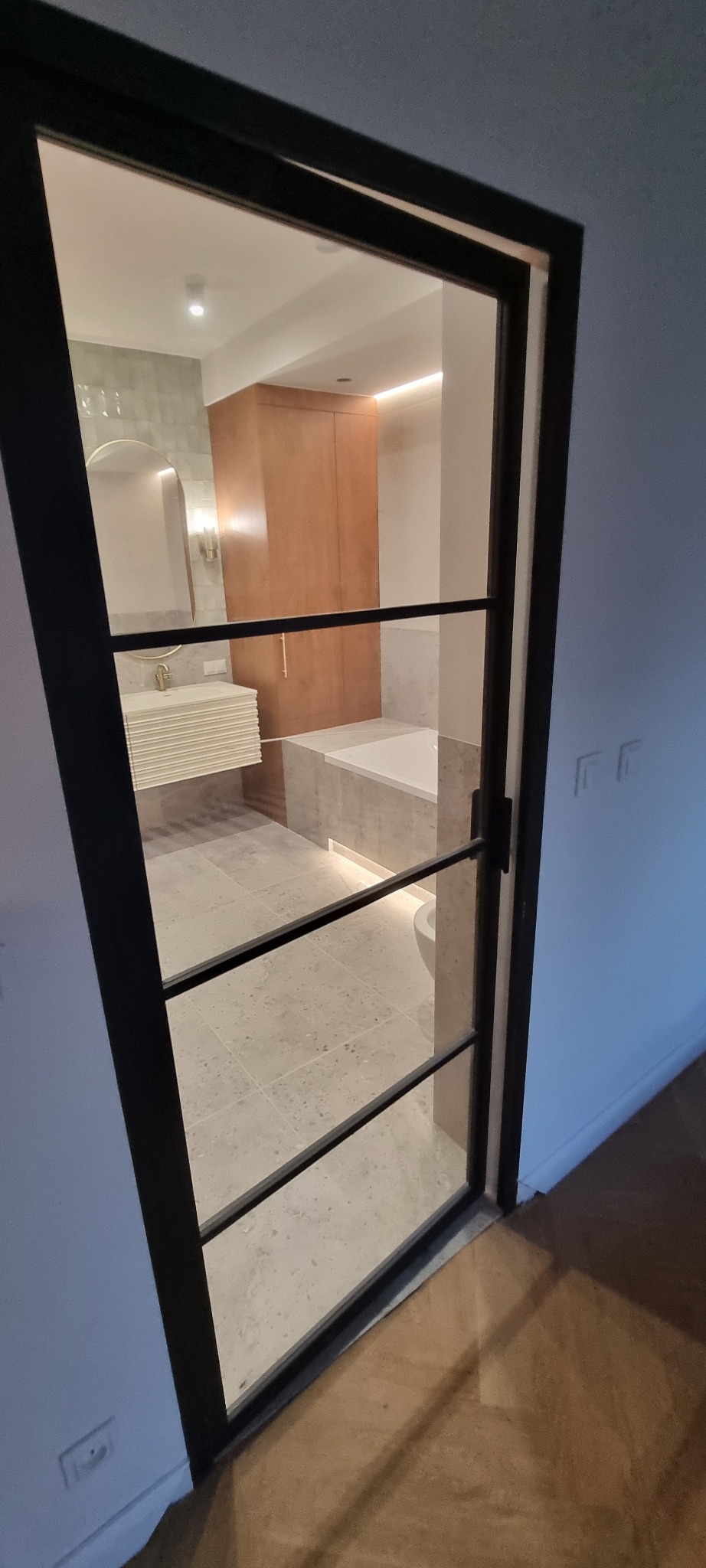 Nowoczesna łazienka widziana przez czarne, przeszklone drzwi z poziomymi szprosami. Widoczna wanna, umywalka i szafka w jasnych barwach.