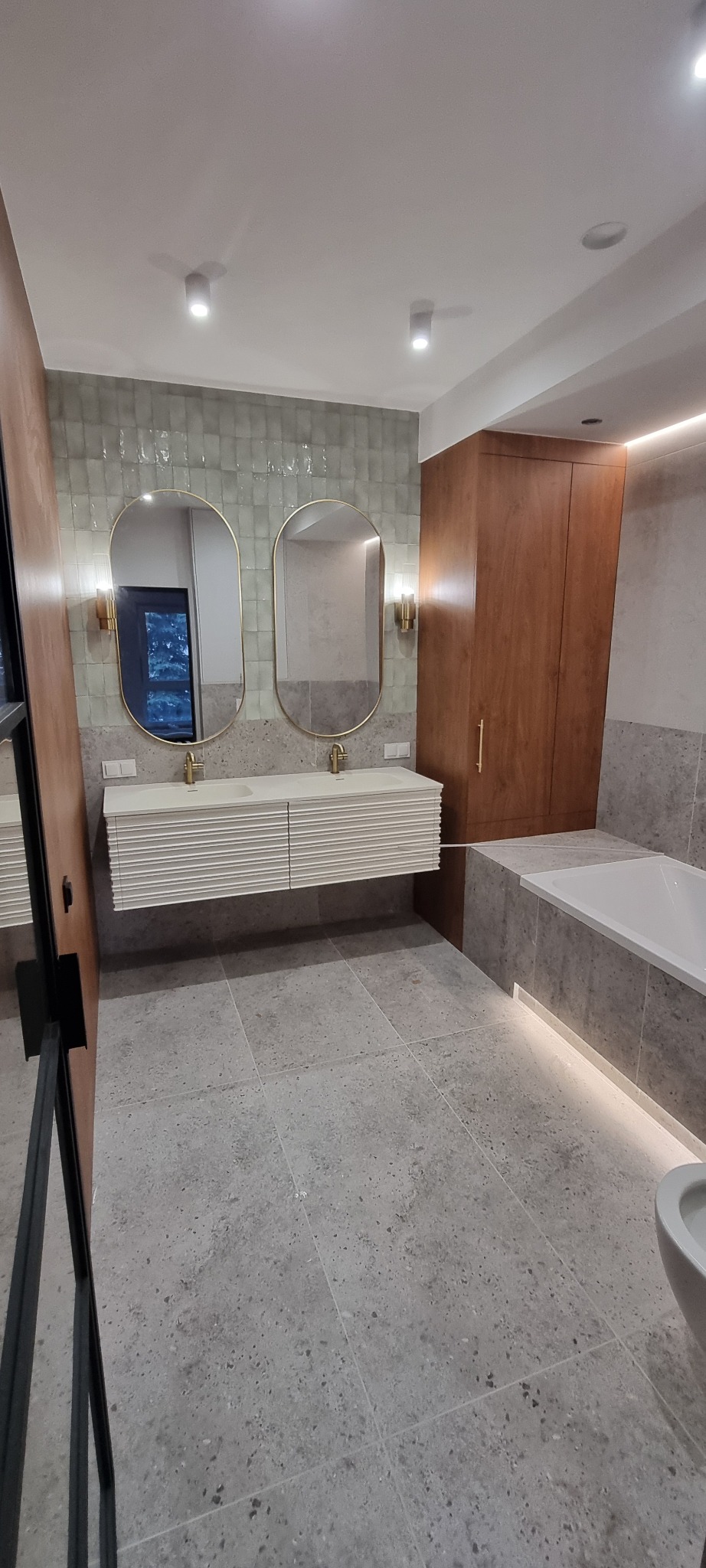 Nowoczesna łazienka z dwoma owalnymi lustrami w złotych ramach nad białą, ryflowaną szafką umywalkową. Szare płytki podłogowe i ścienne, drewniana szafa w zabudowie.