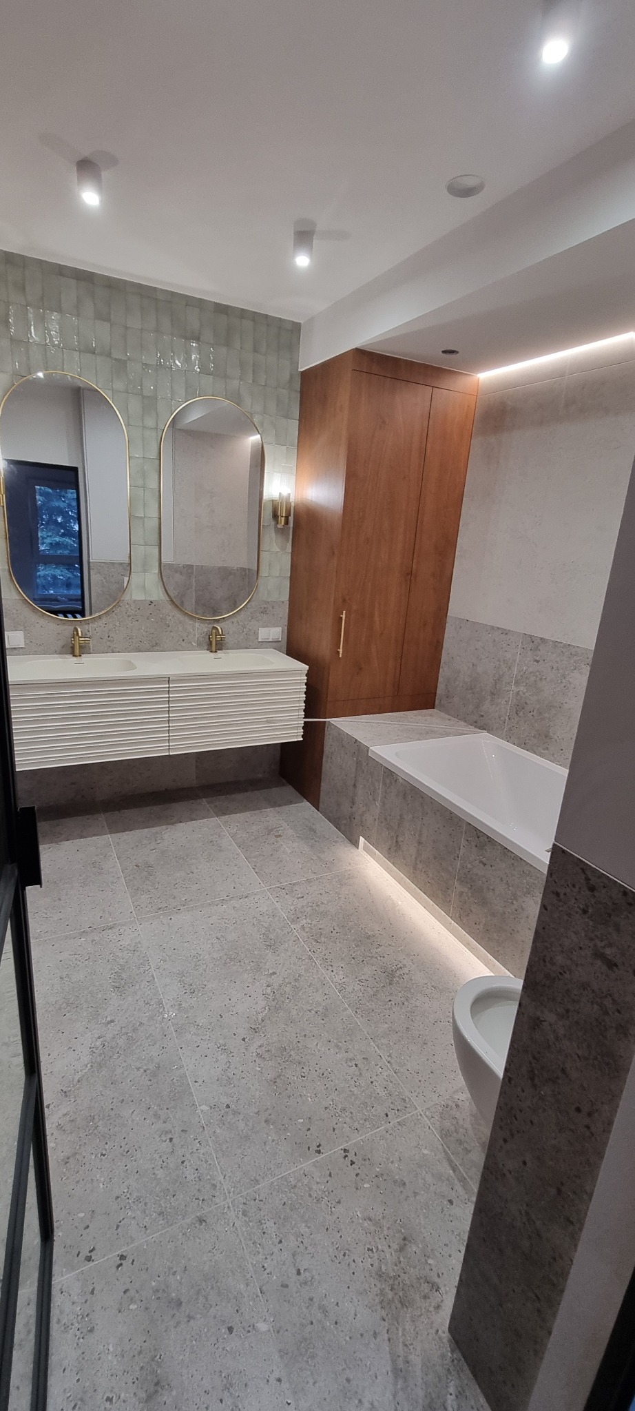 Nowoczesna łazienka z podświetlaną wanną, dwoma owalnymi lustrami w złotych ramach i szarą podłogą z płytek. Uwagę przykuwa drewniana szafa w zabudowie.