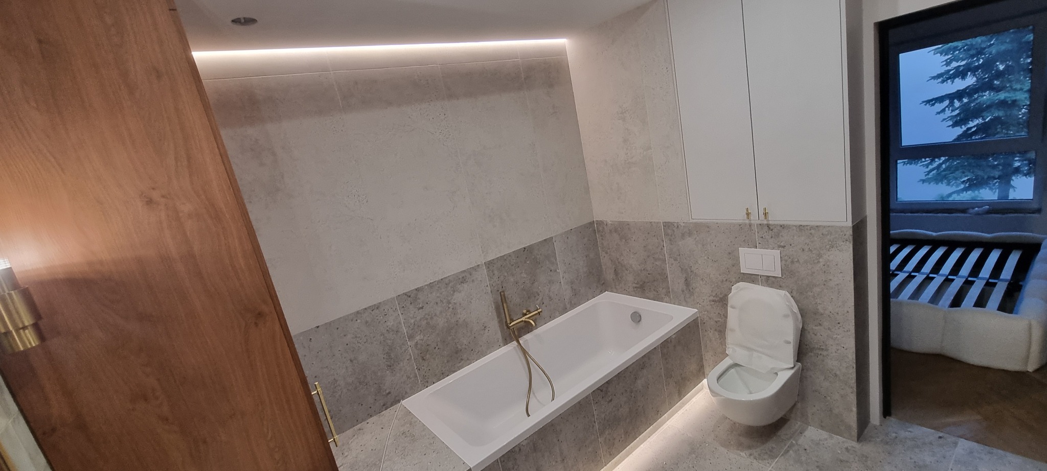 Nowoczesna łazienka z szarą mozaiką, białą wanną z złotą armaturą i podświetleniem LED. Widok na pokój przez otwarte drzwi.