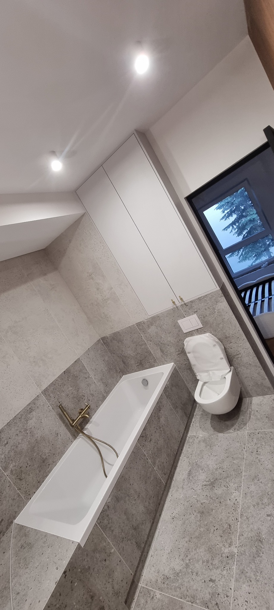 Nowoczesna łazienka z szarymi płytkami, białą wanną i toaletą podwieszaną. Złota armatura dodaje elegancji. Minimalistyczne szafki wiszące.