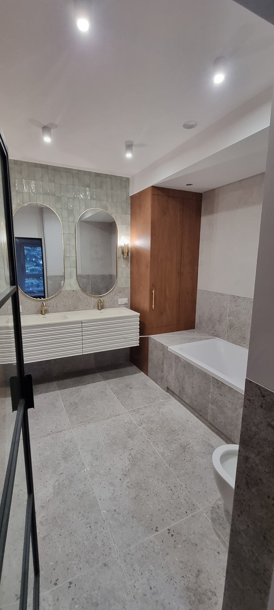 Nowoczesna łazienka z owalnymi lustrami w złotych ramach, białą umywalką z szafką, szarą terakotą i drewnianą szafką. Widoczna wanna i fragment kabiny prysznicowej.