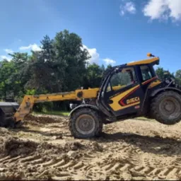 Żółto-czarna ładowarka teleskopowa Dieci z pełną łyżką unosi się na nierównym, piaszczystym terenie budowy, z widocznymi śladami opon i lasem w tle pod błękitnym niebem.