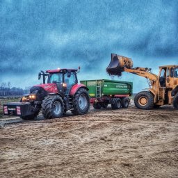 Firma Handlowo Usługowa - Żółta ładowarka czołowa marki Stalowa Wola nabiera ziemię i wsypuje do zielonej przyczepy Pronar Turbo, ciągniętej przez czerwony traktor Case IH na piaszczystym terenie.