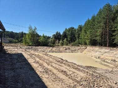 Wyrobisko wypełnione wodą, świeżo po pracach ziemnych, widoczne ślady opon maszyn budowlanych, otoczone zielenią drzew i krzewów w słoneczny dzień.