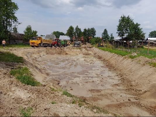 Wykopany, częściowo wypełniony wodą staw w wiejskim otoczeniu, z widocznym ciężkim sprzętem budowlanym i mężczyzną w oddali.