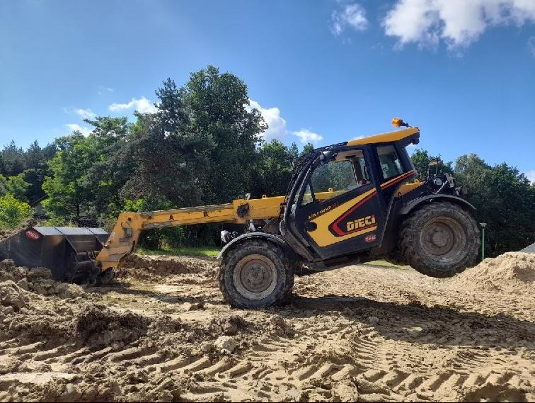 Żółto-czarna ładowarka teleskopowa Dieci z pełną łyżką unosi się na nierównym, piaszczystym terenie budowy, z widocznymi śladami opon i lasem w tle pod błękitnym niebem.