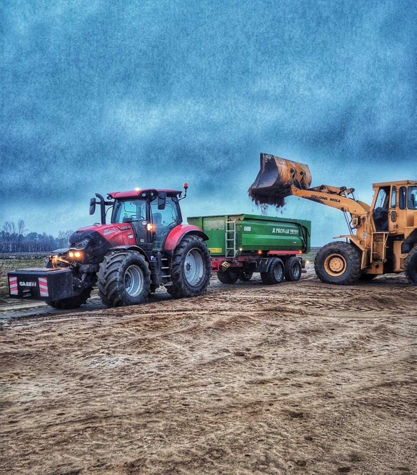 Żółta ładowarka czołowa marki Stalowa Wola nabiera ziemię i wsypuje do zielonej przyczepy Pronar Turbo, ciągniętej przez czerwony traktor Case IH na piaszczystym terenie.