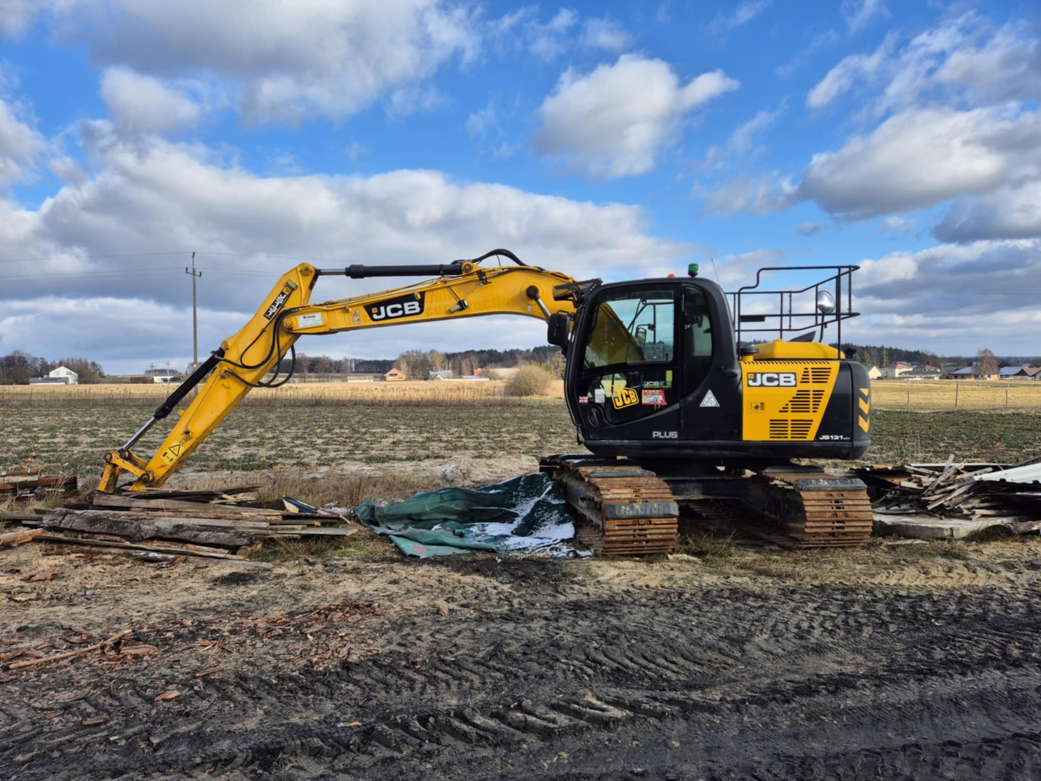 Żółto-czarna koparka JCB na polu z drewnianymi odpadami i śladami gąsienic w ziemi, błękitne niebo z chmurami w tle.
