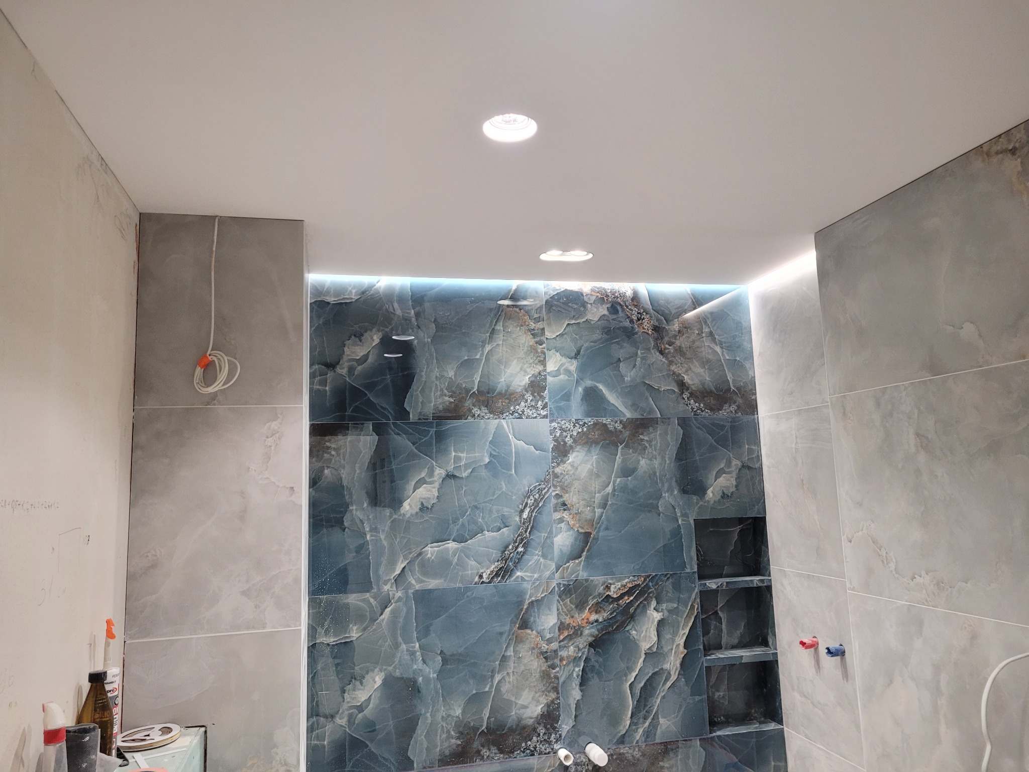 Wykończona łazienka z marmurowymi płytkami w odcieniach szarości i błękitu. Widoczne oświetlenie LED i wnęki w ścianie. Częściowo widoczne przewody elektryczne.