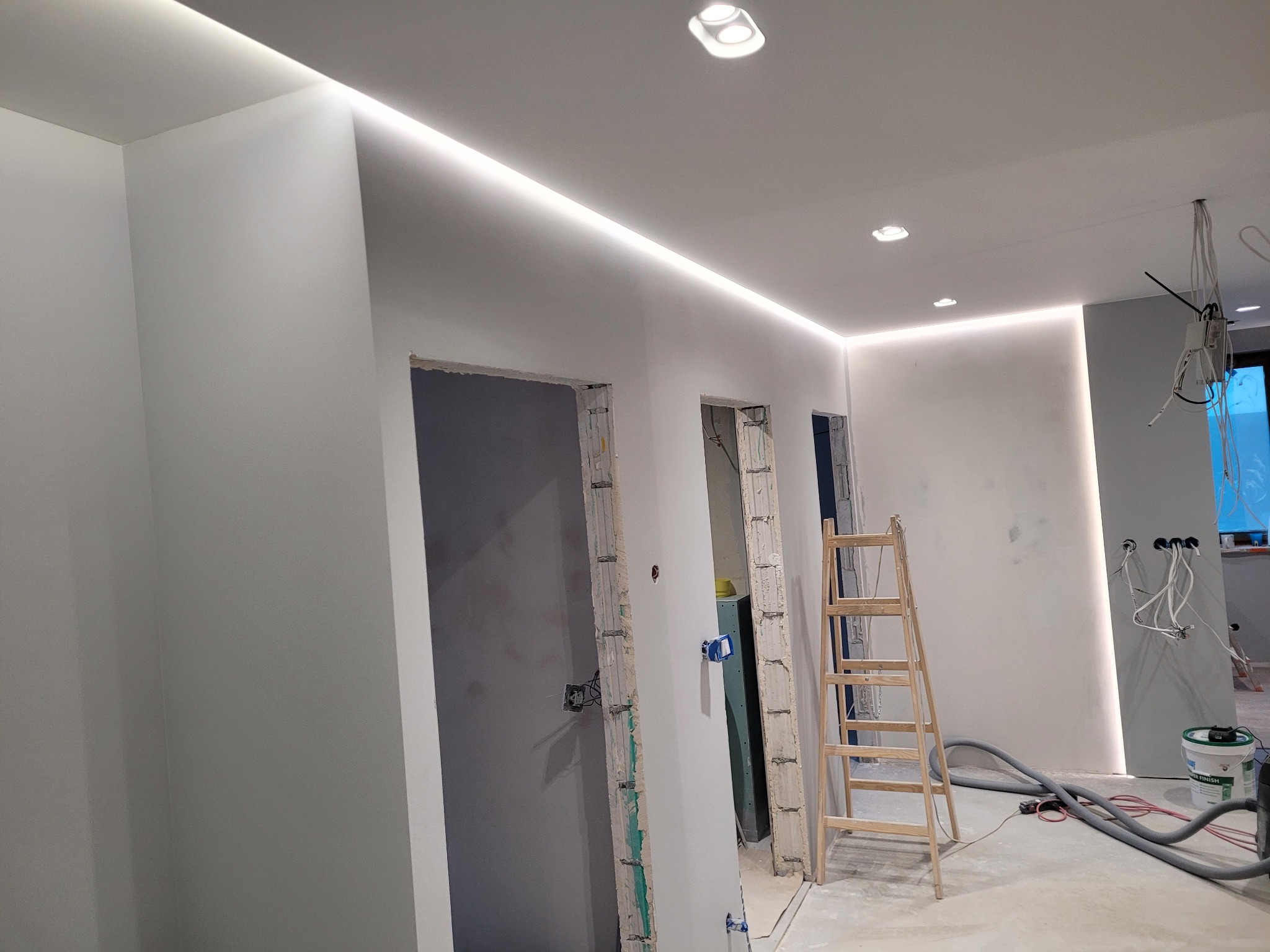 Białe wnętrze w trakcie montażu oświetlenia LED, widoczne listwy LED na suficie i ścianach, drabina oraz przygotowane otwory na gniazdka elektryczne.