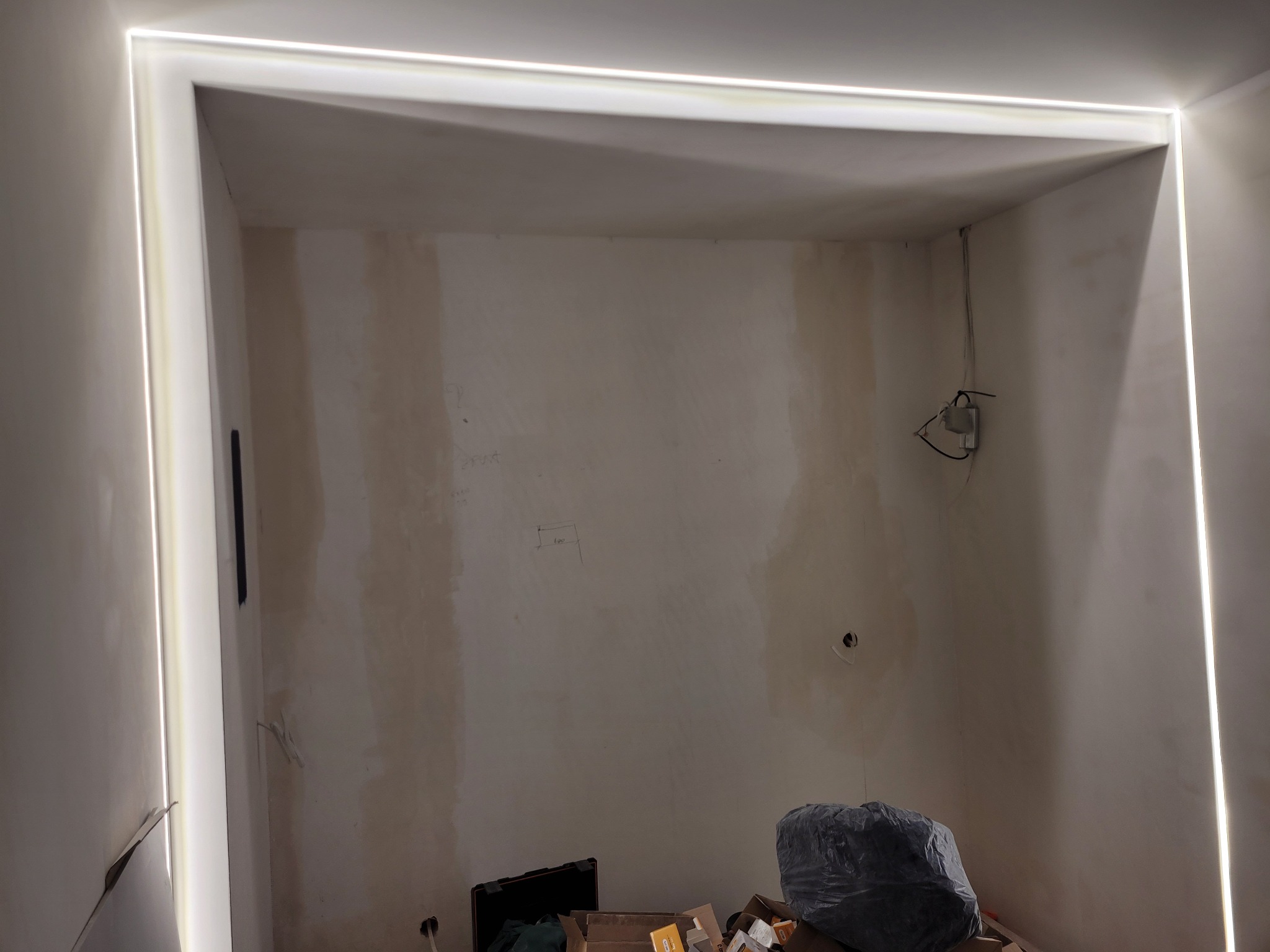 Wnęka z zamontowanym oświetleniem LED wzdłuż krawędzi sufitu i ścian. Widoczne niedoskonałości ścian przed wykończeniem. Na podłodze kartony i worek ze śmieciami.