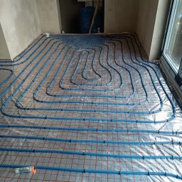 TP OZE INSTAL Tadeusz Pastuszka - Rozłożone niebieskie rury ogrzewania podłogowego na folii z czerwoną siatką, widoczne mocowania i narzędzie, surowe ściany w tle.