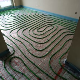 TP OZE INSTAL Tadeusz Pastuszka - Układanie zielonych rur grzewczych na folii izolacyjnej z siatką w nowym budynku, widoczne niebieskie uchwyty mocujące i fragment okna.