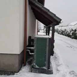 Zewnętrzna jednostka pompy ciepła, zielone lamele, zainstalowana przy ścianie domu, osłonięta drewnianym zadaszeniem, widok w zimowej scenerii, śnieg na ziemi.