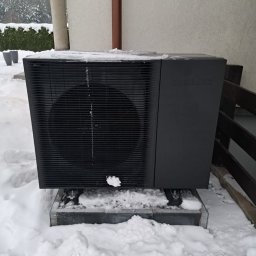 TP OZE INSTAL Tadeusz Pastuszka - Czarna pompa ciepła Samsung, częściowo pokryta śniegiem, stoi na betonowej podstawie w zimowym krajobrazie przy budynku z drewnianym ogrodzeniem.