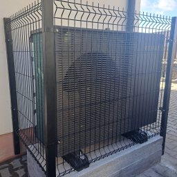 TP OZE INSTAL Tadeusz Pastuszka - Czarna pompa ciepła Samsung zabezpieczona metalowym ogrodzeniem panelowym na betonowej podstawie, widoczna ściana budynku i fragment kostki brukowej.