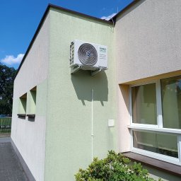 TP OZE INSTAL Tadeusz Pastuszka - Zewnętrzny klimatyzator Kaisai Eco zamontowany na ścianie budynku o dwukolorowej elewacji, widoczny fragment okna i zieleń krzewu, błękitne niebo w tle.