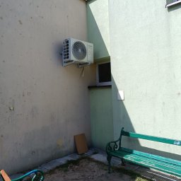TP OZE INSTAL Tadeusz Pastuszka - Zamontowana jednostka zewnętrzna klimatyzatora Kaisai Eco na ścianie budynku, widoczne dwie ławki parkowe w tle.