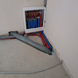 TP OZE INSTAL Tadeusz Pastuszka - Rozdzielacz ogrzewania podłogowego z niebieskimi i czerwonymi rurami w białej obudowie, szare rury kanalizacyjne na surowej podłodze. Instalacja w trakcie realizacji.