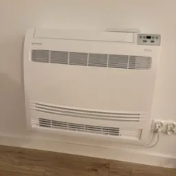 Biały klimakonwektor przypodłogowy w Miedźnej, zamontowany na ścianie. Widoczne kratki wentylacyjne i panel sterowania. Gniazdka elektryczne w pobliżu.