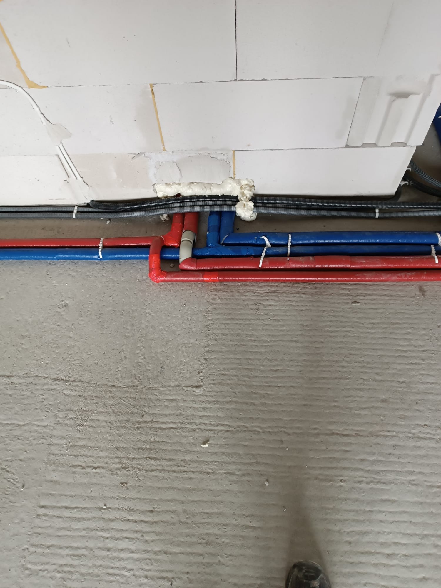 Czerwone i niebieskie rury wodne, zabezpieczone opaskami, biegnące wzdłuż ściany z bloczków, z widoczną pianką montażową i przewodami elektrycznymi.