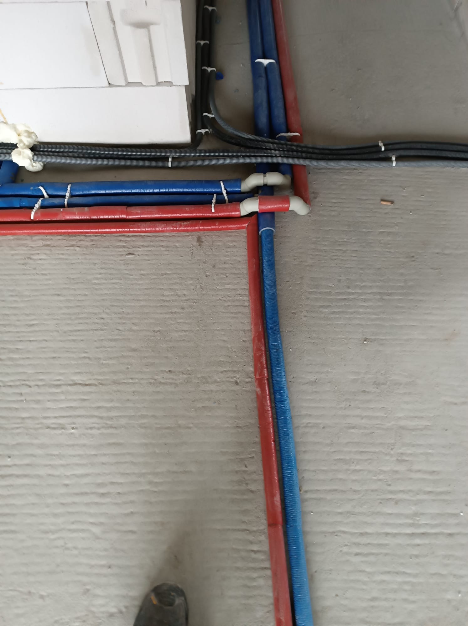 Czerwone i niebieskie rury instalacji wodnej ułożone na szarym, surowym podłożu betonowym, zabezpieczone białymi opaskami zaciskowymi, z widocznymi fragmentami instalacji elektrycznej w peszlu...