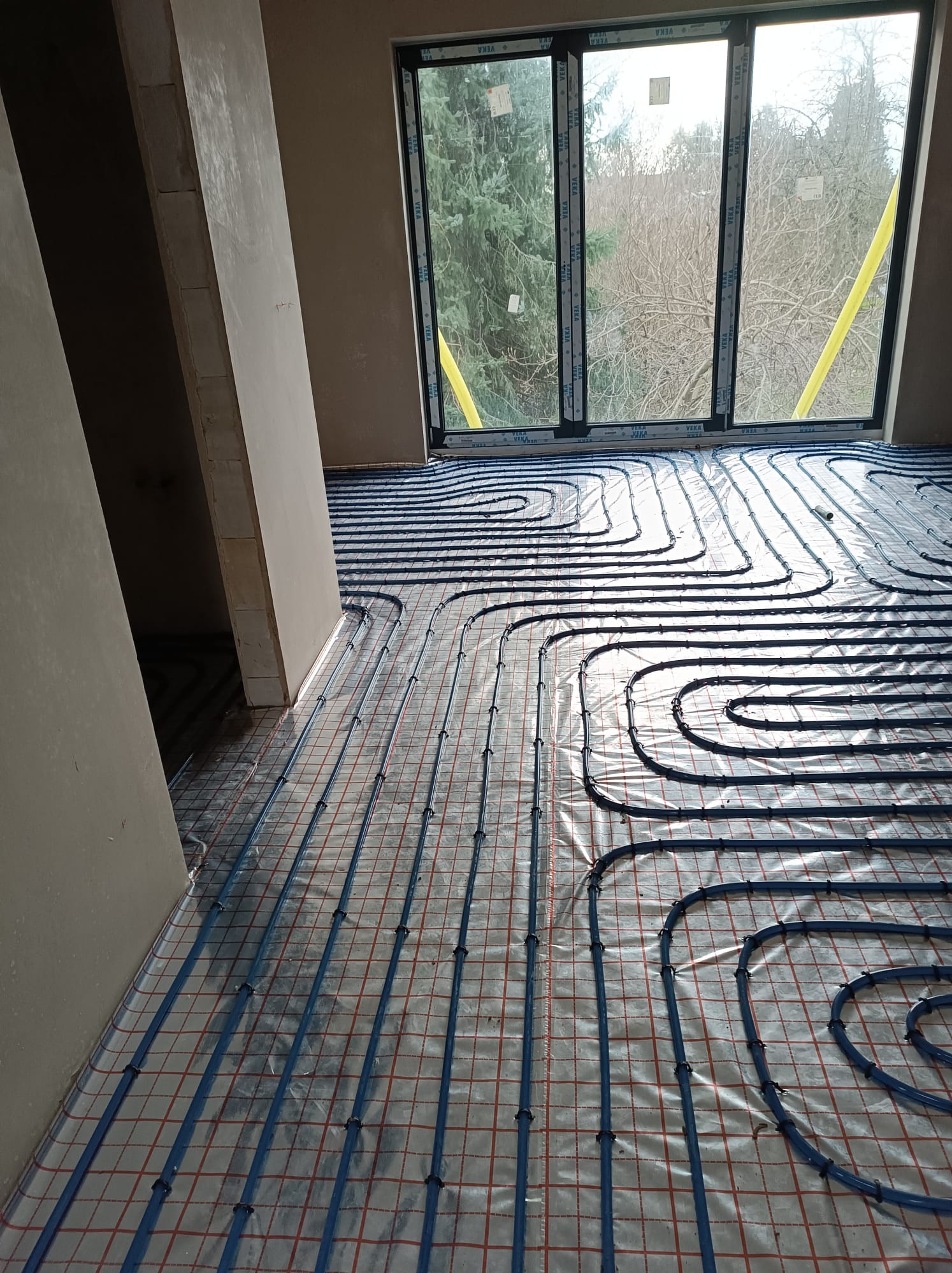 Niebieskie rury ogrzewania podłogowego rozłożone na folii z czerwoną siatką, widoczne okno z żółtymi taśmami zabezpieczającymi i surowe ściany w tle.