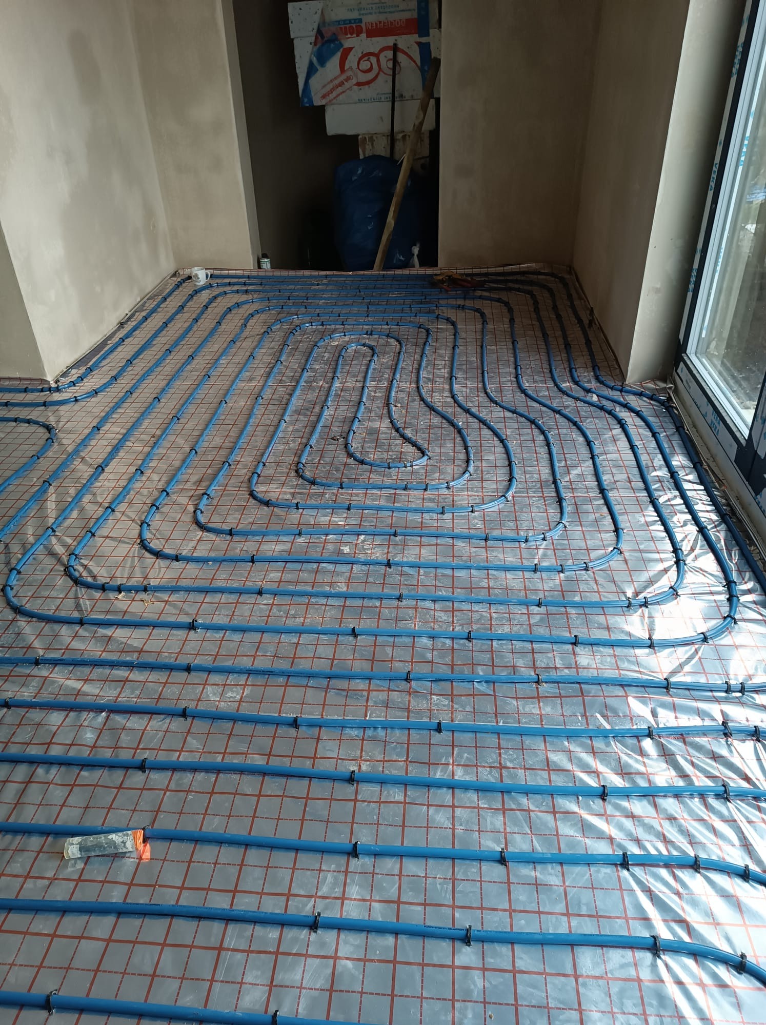 Rozłożone niebieskie rury ogrzewania podłogowego na folii z czerwoną siatką, widoczne mocowania i narzędzie, surowe ściany w tle.
