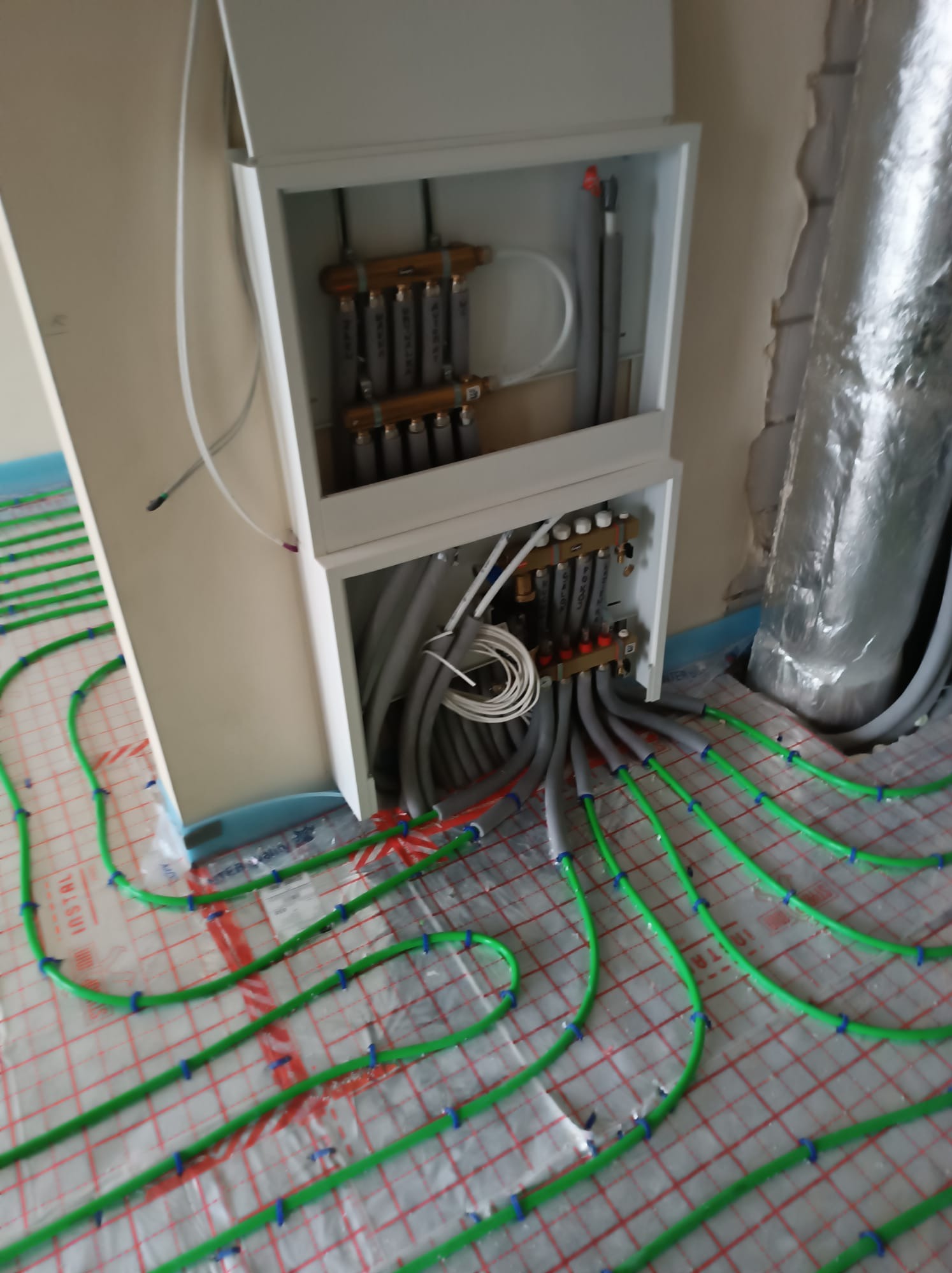 Rozdzielacz ogrzewania podłogowego w białej szafce, z zielonymi rurami ułożonymi na folii izolacyjnej z czerwoną siatką, widoczne szare rury doprowadzające wodę.
