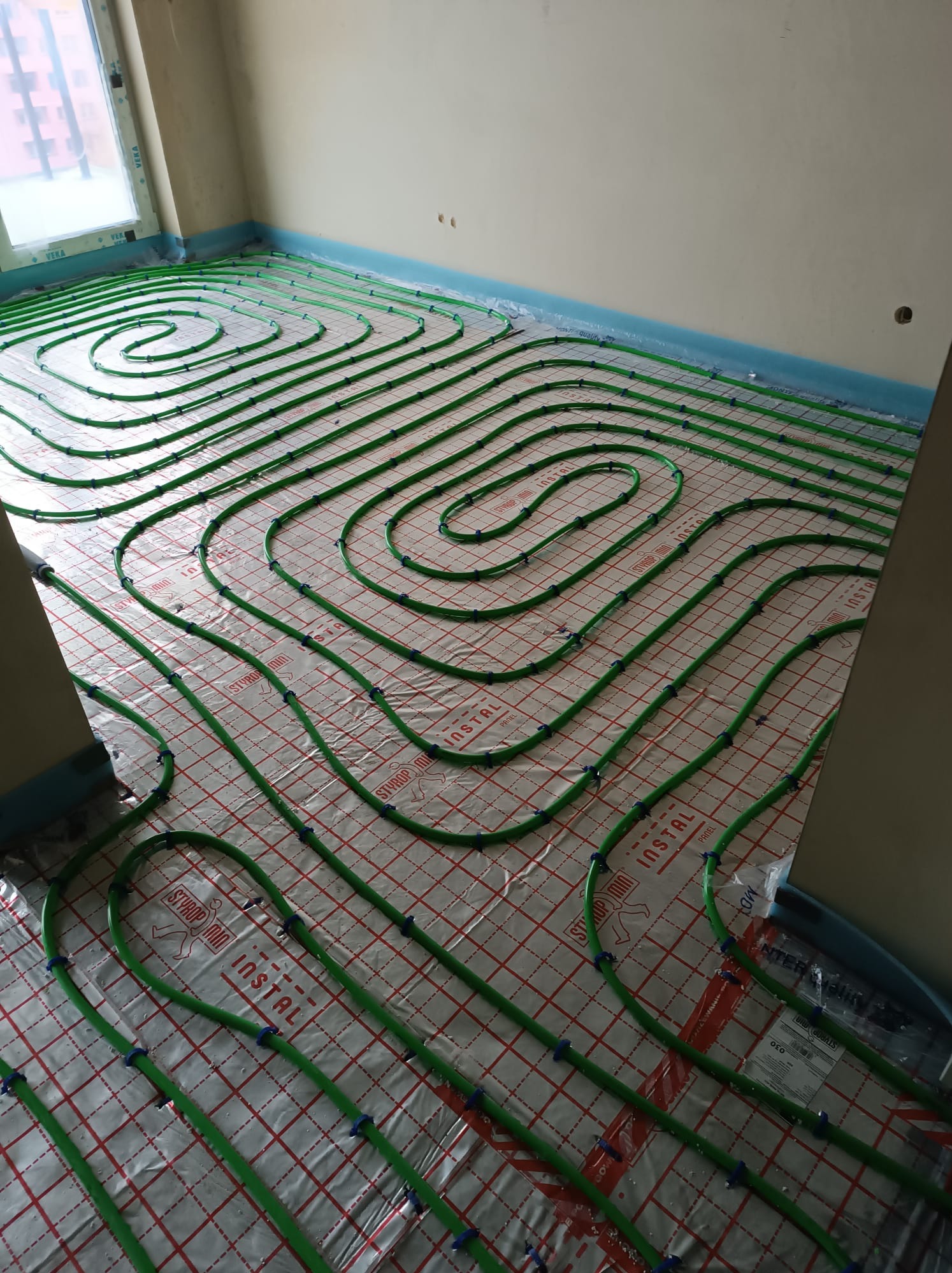 Układanie zielonych rur grzewczych na folii izolacyjnej z siatką w nowym budynku, widoczne niebieskie uchwyty mocujące i fragment okna.