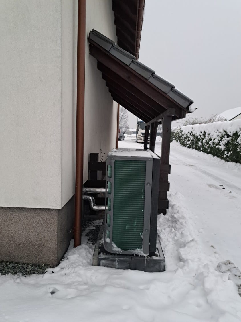Zewnętrzna jednostka pompy ciepła, zielone lamele, zainstalowana przy ścianie domu, osłonięta drewnianym zadaszeniem, widok w zimowej scenerii, śnieg na ziemi.