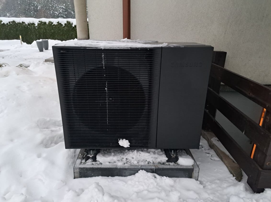 Czarna pompa ciepła Samsung, częściowo pokryta śniegiem, stoi na betonowej podstawie w zimowym krajobrazie przy budynku z drewnianym ogrodzeniem.