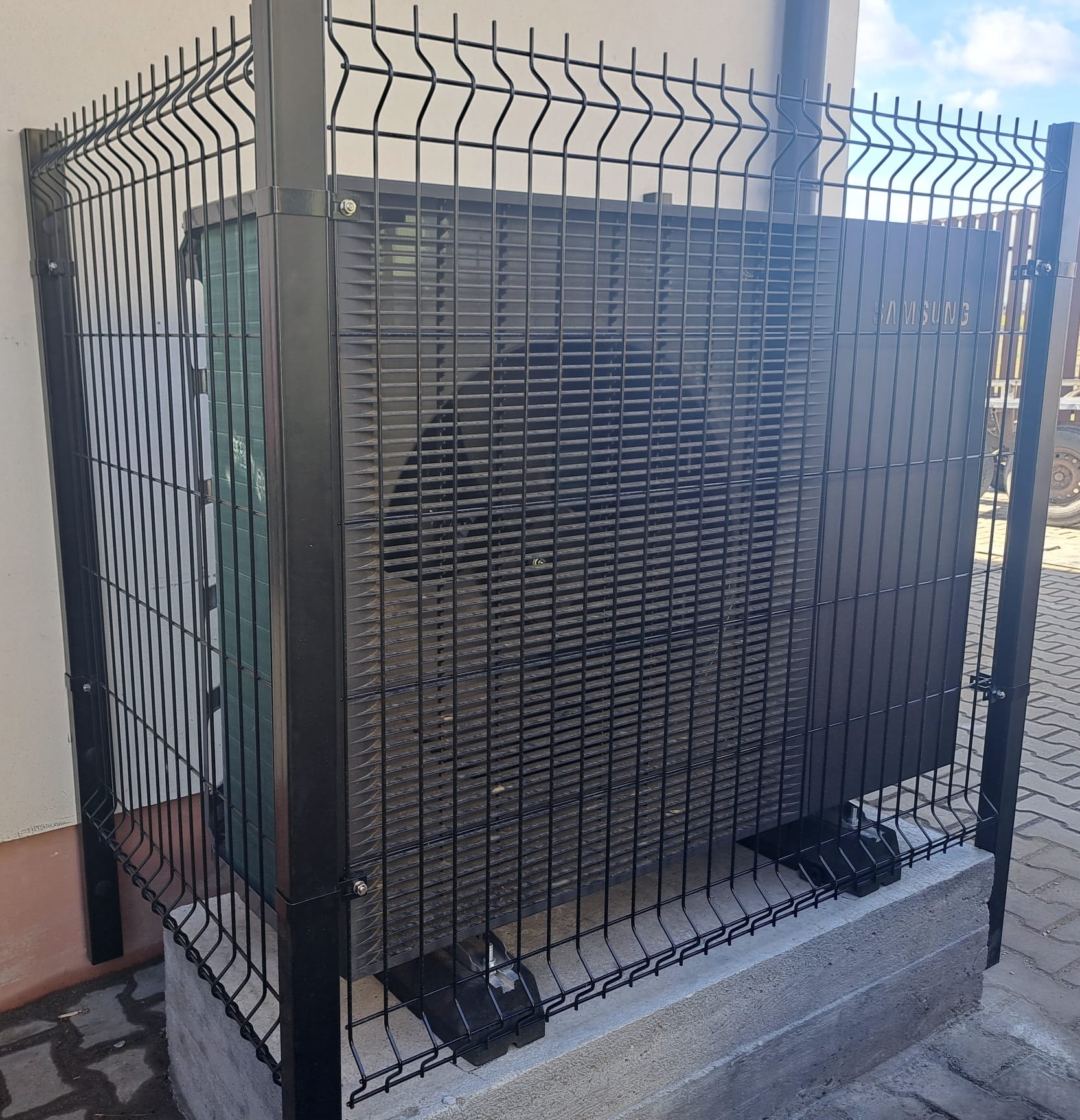 Czarna pompa ciepła Samsung zabezpieczona metalowym ogrodzeniem panelowym na betonowej podstawie, widoczna ściana budynku i fragment kostki brukowej.