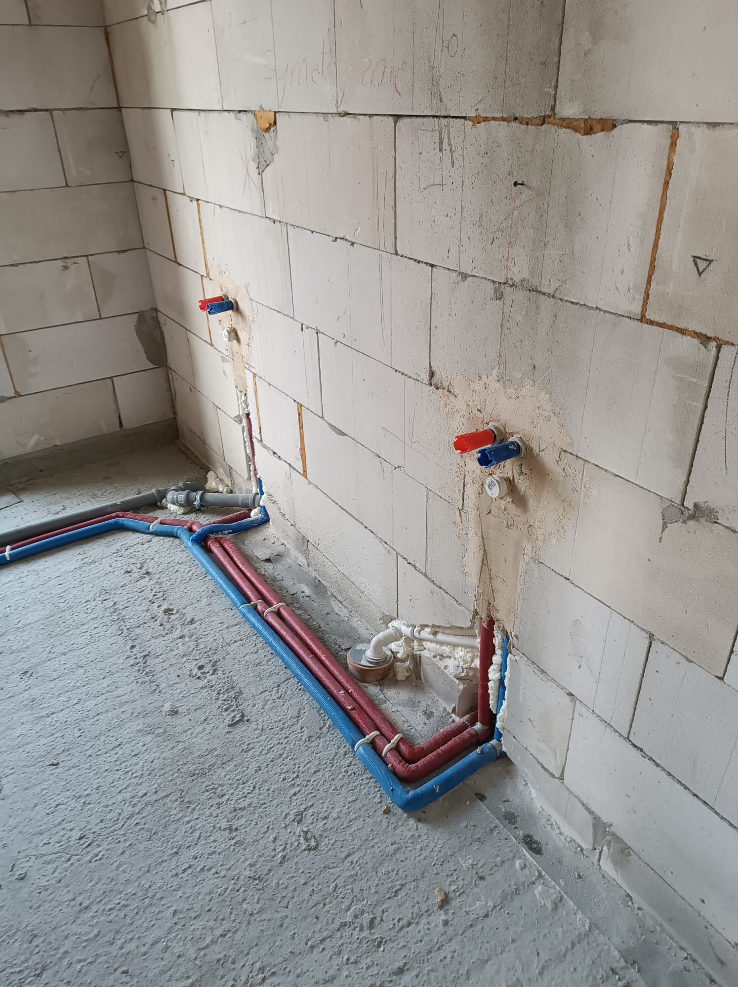 Surowy stan: czerwono-niebieskie rury instalacji wodnej prowadzone po szarym, betonowym podłożu i przymocowane do ściany z białych bloczków, widoczne zawory z czerwoną i niebieską nakładką.