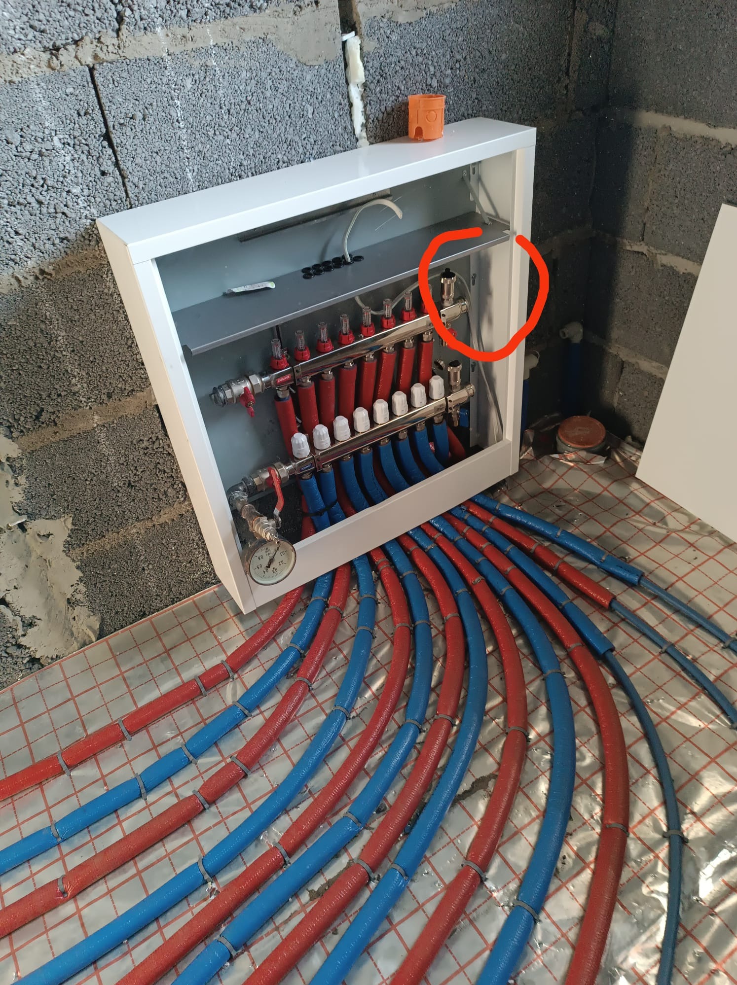 Rozdzielacz ogrzewania podłogowego z czerwonymi i niebieskimi rurami na folii izolacyjnej, widoczne przyłącza i manometr, surowy mur z betonu komórkowego w tle.