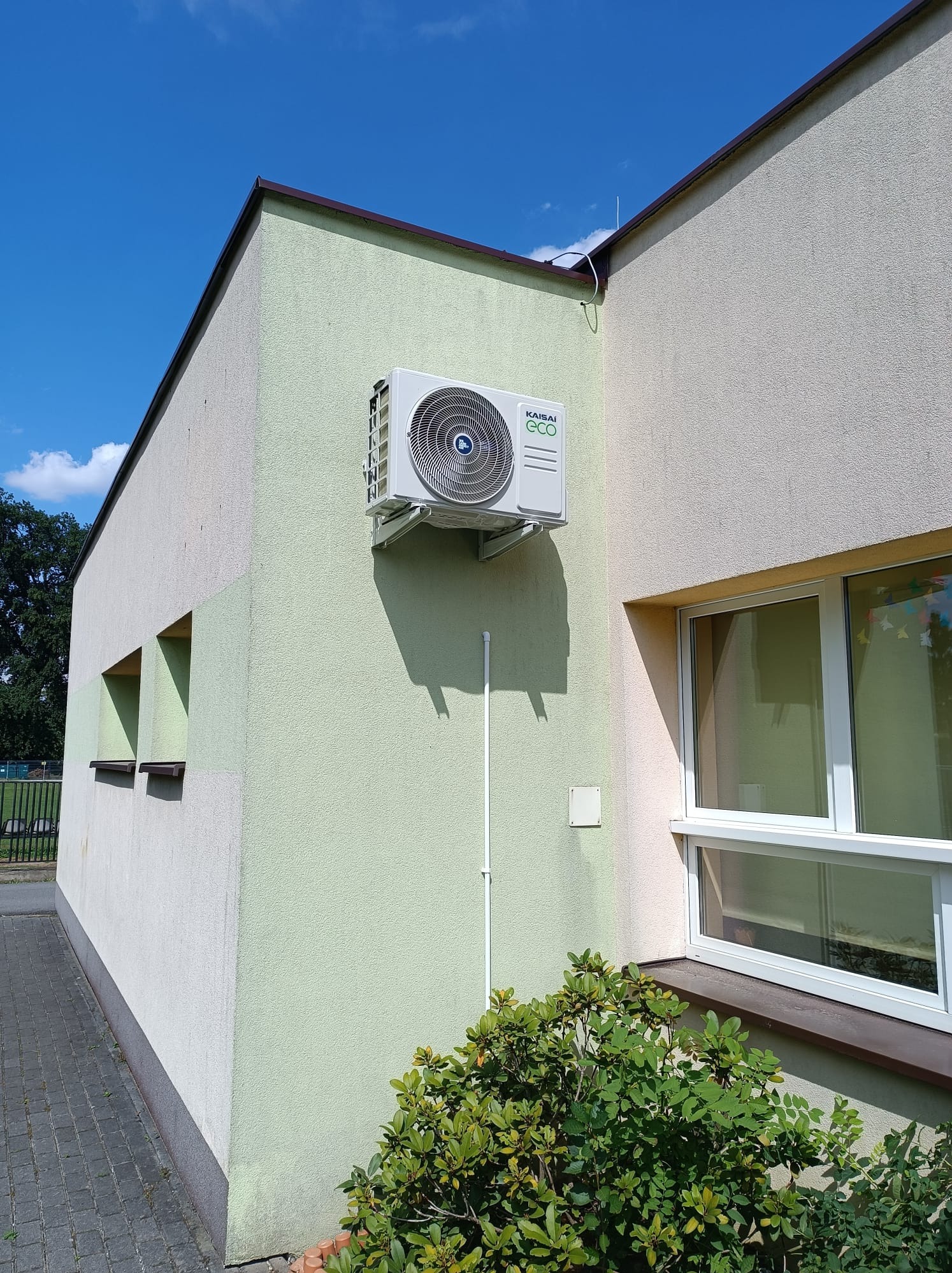 Zewnętrzny klimatyzator Kaisai Eco zamontowany na ścianie budynku o dwukolorowej elewacji, widoczny fragment okna i zieleń krzewu, błękitne niebo w tle.