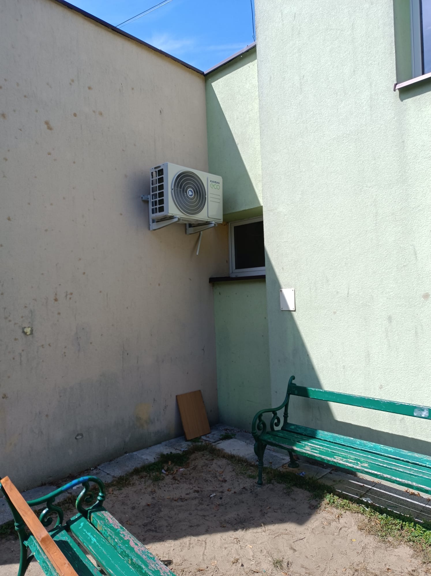 Zamontowana jednostka zewnętrzna klimatyzatora Kaisai Eco na ścianie budynku, widoczne dwie ławki parkowe w tle.