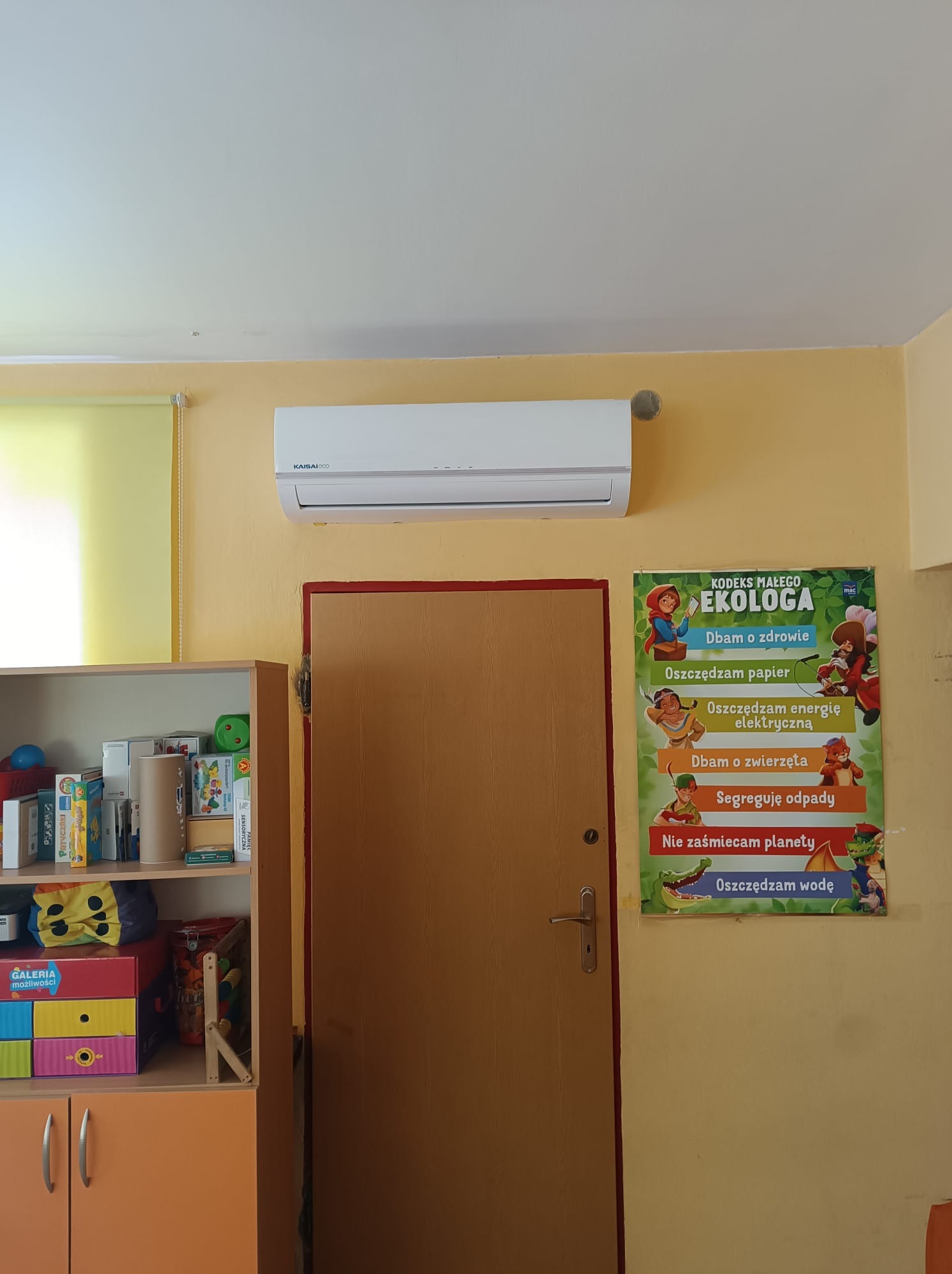 Klimatyzator Kaisai zamontowany na żółtej ścianie w przedszkolu, obok drzwi i plakatu z zasadami ekologii dla dzieci.