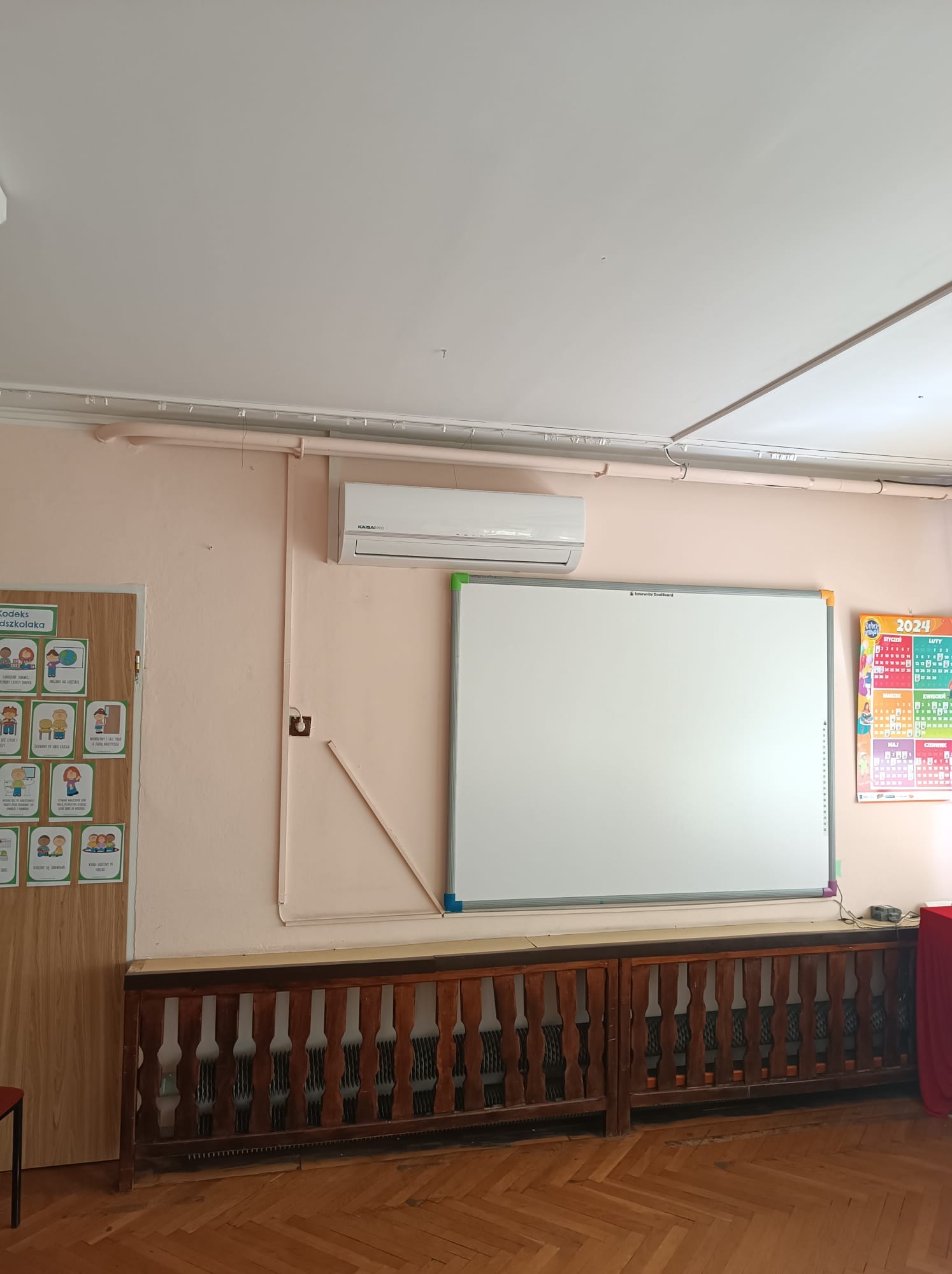 Wnętrze klasy z zamontowaną klimatyzacją Kaisai na ścianie, białą tablicą interaktywną, kalendarzem ściennym na rok 2024 oraz drewnianym, ażurowym obudowaniem grzejnika.