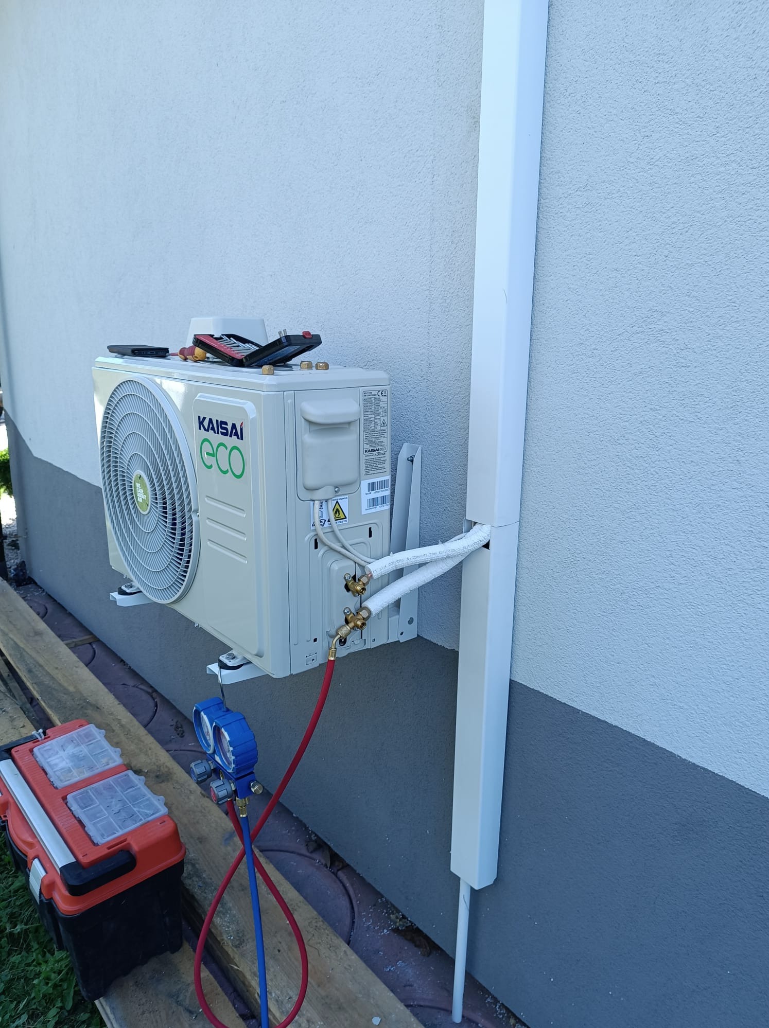 Klimatyzator Kaisai ECO zamontowany na ścianie budynku, widoczne podłączenia rur i narzędzia w skrzynce obok, w trakcie montażu lub serwisu.