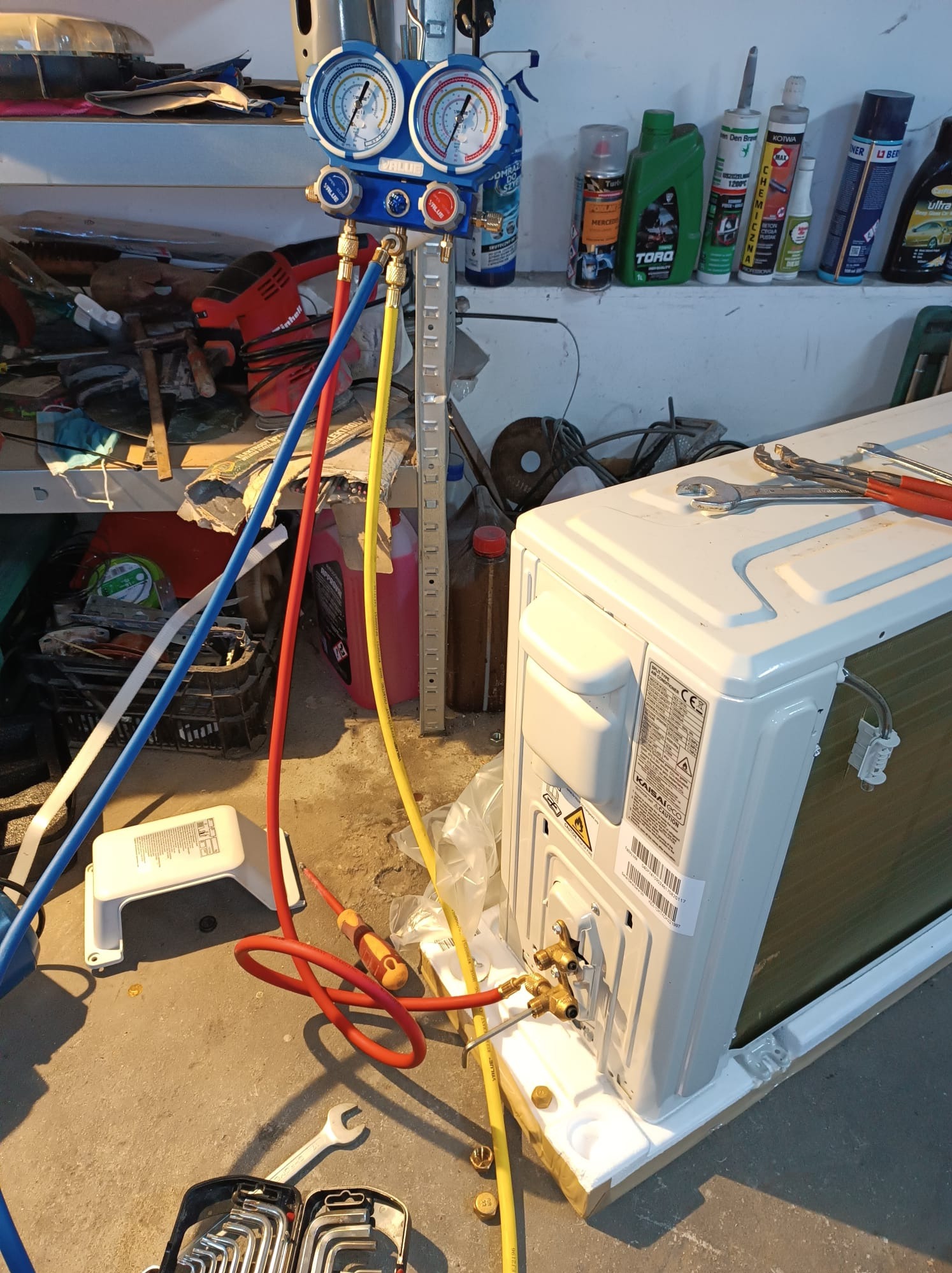 Podłączenie manometru serwisowego do klimatyzatora typu split podczas instalacji; widoczne przewody pomiarowe w kolorach niebieskim, czerwonym i żółtym, narzędzia oraz butelki chemii warsztatowej...
